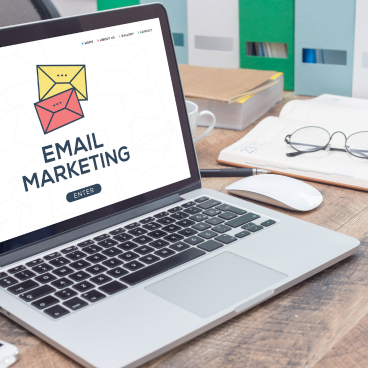 Laptop displaying an email marketing concept with colorful envelope icons, placed on a modern office desk with notebook, glasses, and stationery